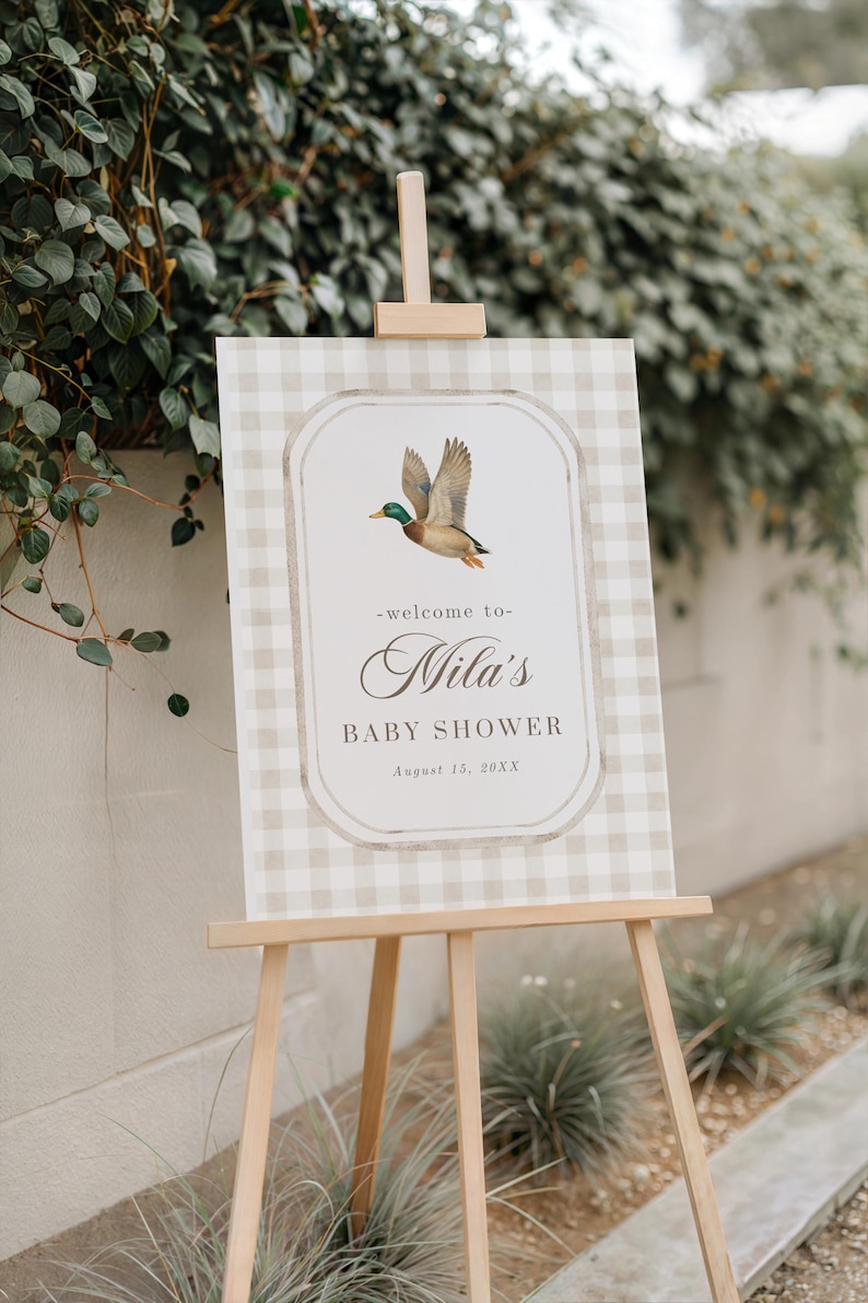 May include: A baby shower sign on a wooden easel. The sign has a beige and white gingham pattern with a central oval featuring a flying duck illustration. Text reads "-welcome to- Mila's BABY SHOWER August 15, 20XX."