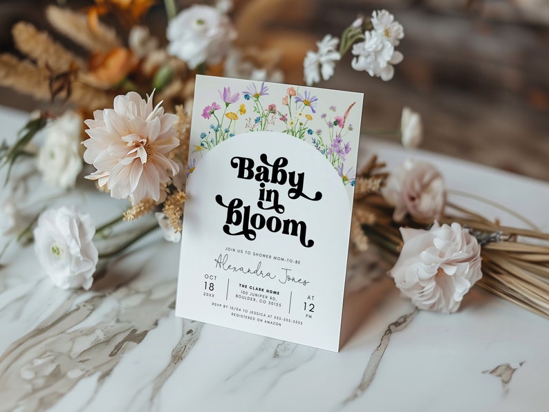 May include: A white baby shower invitation with a floral border and the text "Baby in Bloom" in a black retro font. The invitation includes the date, time, and location of the shower for Alexandra Jones.