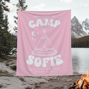 May include: Pink fabric banner with white text: "WELCOME TO CAMP SOFIE". Features a teepee illustration, moon, and stars. Includes "SOFIE'S 1ST BIRTHDAY OCTOBER 23, 20XX". Banner is hanging outdoors near a lake and mountains.