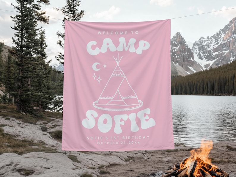 May include: Pink fabric banner with white text: "WELCOME TO CAMP SOFIE". Features a teepee illustration, moon, and stars. Includes "SOFIE'S 1ST BIRTHDAY OCTOBER 23, 20XX". Banner is hanging outdoors near a lake and mountains.