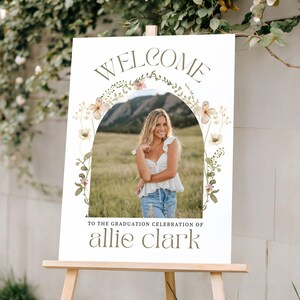 Pu&ograve; includere: Un cartello di benvenuto per una festa di laurea su un cavalletto di legno. Il cartello presenta una foto di una persona con un top bianco e jeans, incorniciata da un arco floreale. Il testo recita "WELCOME" e "TO THE GRADUATION CELEBRATION OF allie clark."