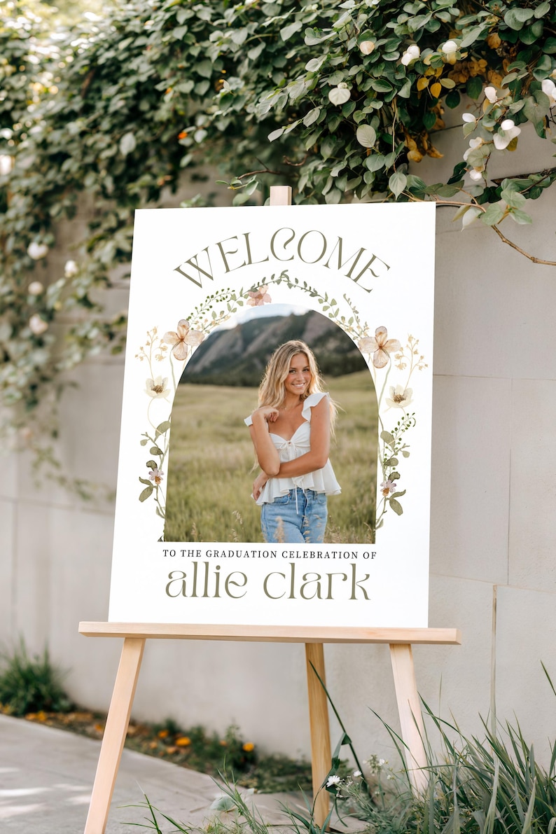 Pu&ograve; includere: Un cartello di benvenuto per una festa di laurea su un cavalletto di legno. Il cartello presenta una foto di una persona con un top bianco e jeans, incorniciata da un arco floreale. Il testo recita "WELCOME" e "TO THE GRADUATION CELEBRATION OF allie clark."