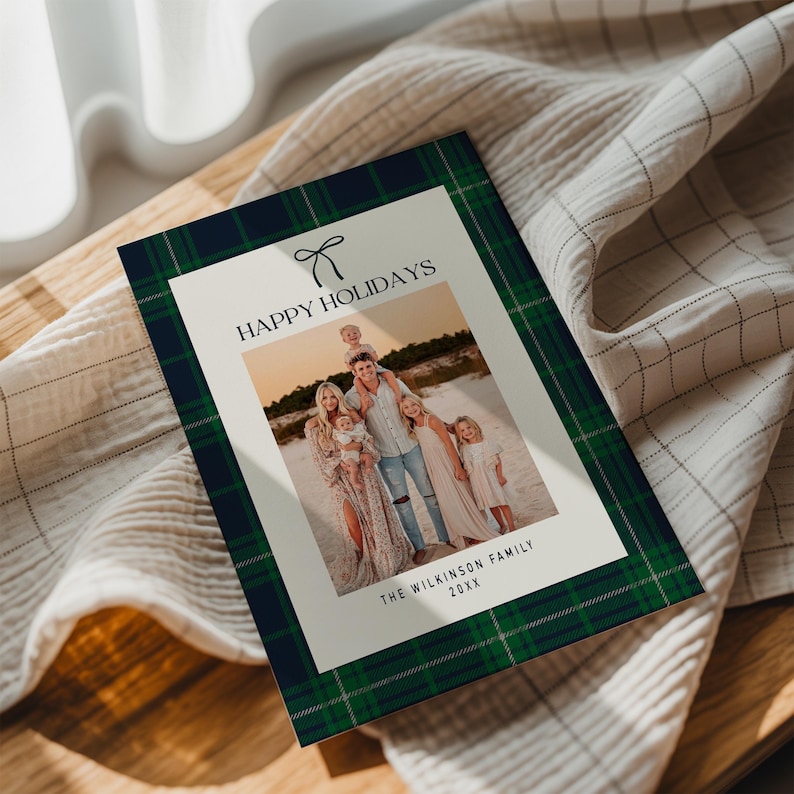 May include: A holiday card with a family photo, framed by a green and navy plaid pattern. The card reads "HAPPY HOLIDAYS" with a bow graphic and "THE WILKINSON FAMILY 20XX" at the bottom. The card is on a light-colored fabric.