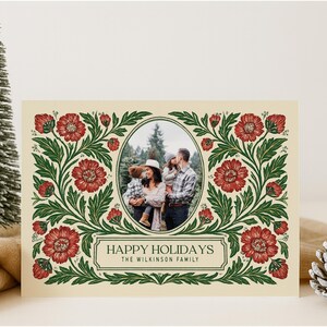 May include: Christmas card featuring a family photograph within an oval frame, framed by a floral pattern of red flowers and green foliage. The text reads "HAPPY HOLIDAYS" and the family name. A small Christmas tree and pine cone are in the background.