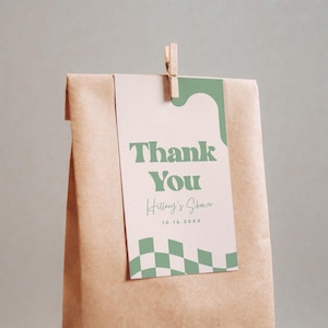 May include: A brown paper bag with a rectangular tag attached by a wooden clothespin. The tag features a light pink background, sage green wavy accents, a green checkered pattern, and the text Thank You, Hillary's Shower, and 10.16.20XX.