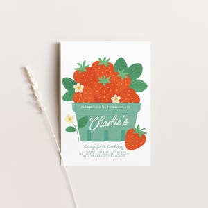 May include: A white invitation card with a berry theme. It features a green basket overflowing with red strawberries, green leaves, and white flowers. The text "PLEASE JOIN US TO CELEBRATE" is above the name "Charlie's". The text "berry first birthday" is below.