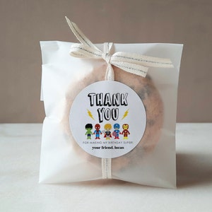 May include: A white thank you favor bag with a round sticker featuring a group of superhero characters. The sticker says "Thank You" with lightning bolts and "For making my birthday super! Your friend, Lucas".