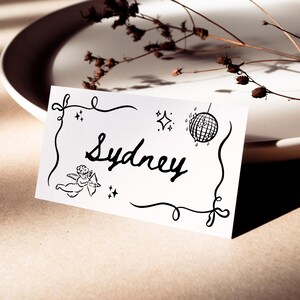 May include: A white place card with the name "Sydney" in black script, framed by a decorative black border. The card features illustrations of a disco ball, stars, and a cherub. The card is placed on a white plate.