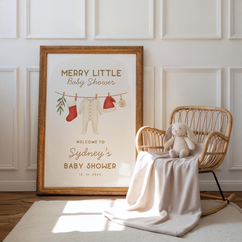 May include: A framed baby shower sign with gold trim, displaying the text "Merry Little Baby Shower" and "Welcome to Sydney's Baby Shower." The sign features illustrations of baby clothes and a Christmas theme. A wicker rocking chair with a stuffed bunny and blanket is next to the sign.