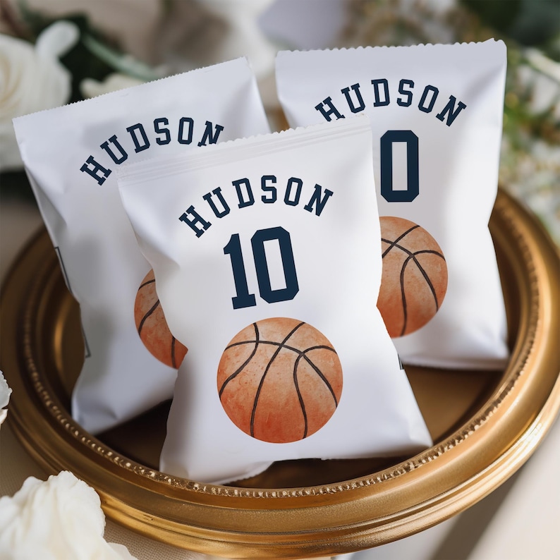 May include: Three white favor bags with a basketball theme. The bags have the name "HUDSON" printed on them, along with a basketball and a number. The numbers are 0 and 10.