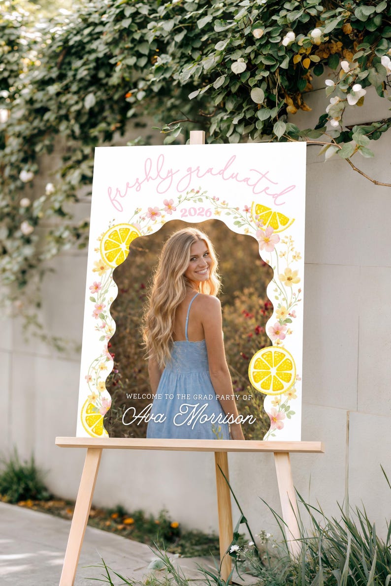 K&ouml;nnte beinhalten: Ein Willkommensschild f&uuml;r eine Abschlussfeier auf einer Holzstaffelei. Das Schild zeigt ein Foto einer Person in einem blauen Kleid, umgeben von einem floralen und Zitronen-Design. Der Text enth&auml;lt "freshly graduated" und "2026" mit einem Namen darunter.