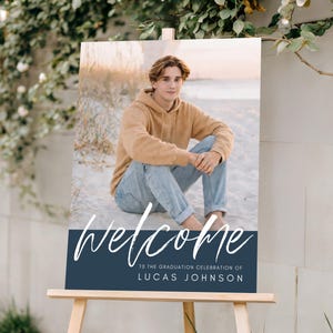 May include: A graduation welcome sign on a wooden easel. The sign features a photo of a person in a tan hoodie and jeans. The sign reads "welcome" in white script, with the text "TO THE GRADUATION CELEBRATION OF LUCAS JOHNSON" below.
