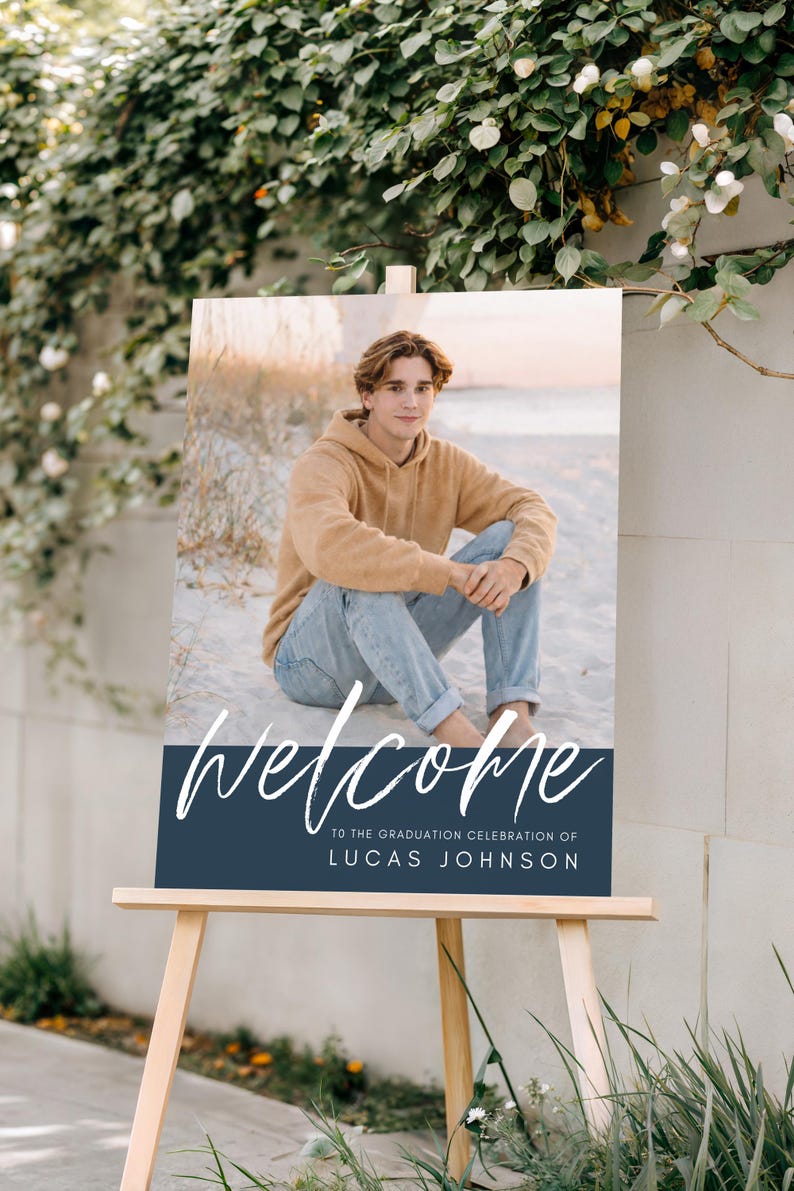 May include: A graduation welcome sign on a wooden easel. The sign features a photo of a person in a tan hoodie and jeans. The sign reads "welcome" in white script, with the text "TO THE GRADUATION CELEBRATION OF LUCAS JOHNSON" below.