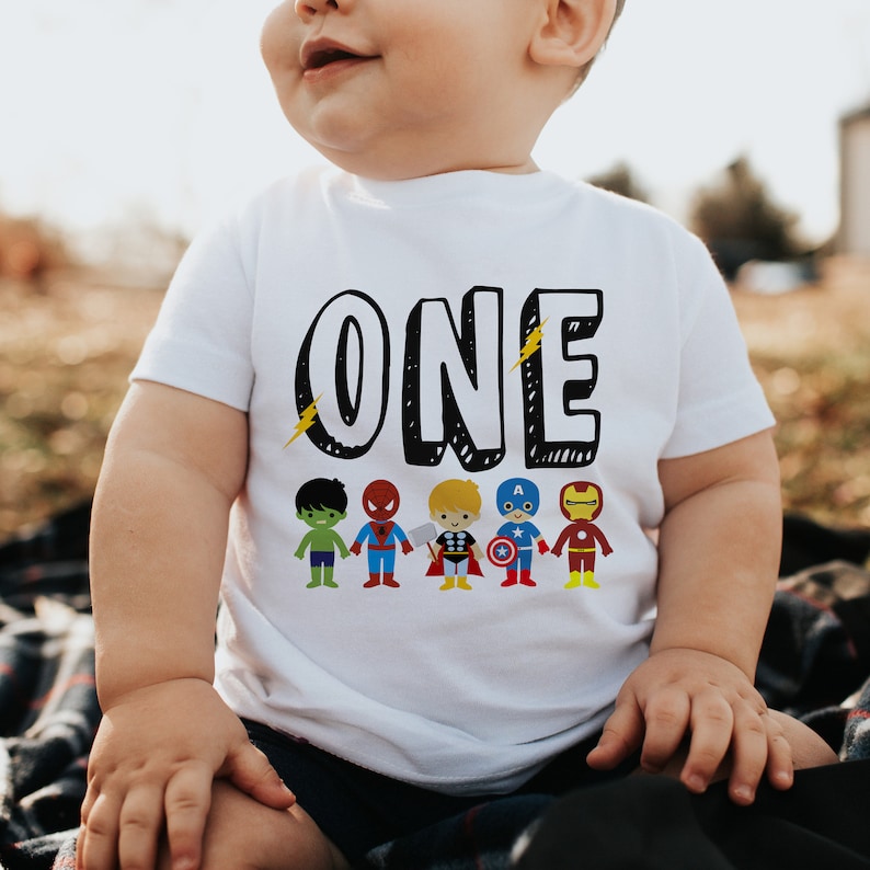 May include: White short-sleeved shirt with the word "ONE" in black outlined letters and a row of superhero characters below. The characters include Hulk, Spiderman, Thor, Captain America, and Iron Man.