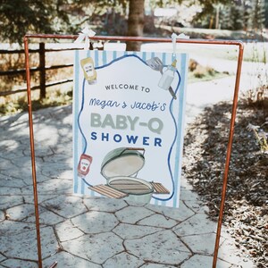 May include: A sign for a baby shower, held outdoors, with a copper frame. The sign reads "Welcome to Megan & Jacob's BABY-Q SHOWER" with illustrations of a grill, condiments, and baby items. The background has blue and white stripes.