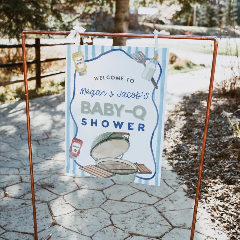 May include: A sign for a baby shower, held outdoors, with a copper frame. The sign reads "Welcome to Megan & Jacob's BABY-Q SHOWER" with illustrations of a grill, condiments, and baby items. The background has blue and white stripes.
