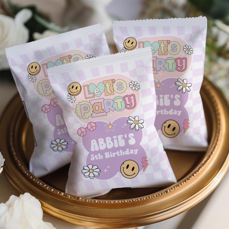 May include: Purple and white party favor bags with a checkered pattern. The bags have a colorful "Let's Party" design with a smiley face, flowers, and stars. The text "Abbie's 5th Birthday" is printed on the bag.