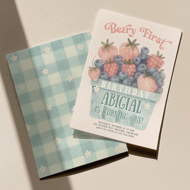 May include: A birthday invitation with a watercolor illustration of berries in a basket. The text "Berry First" and "Abigial is turning one!" are displayed in a whimsical font. The invitation is paired with a light blue gingham patterned envelope.