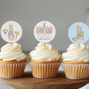 May include: Three cupcakes with white frosting and blue and white Peter Rabbit themed cupcake toppers. The toppers feature a rabbit with a blue jacket and a carrot. One topper says "Marissa's Baby Shower".