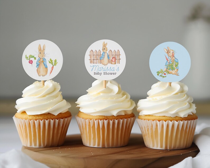 May include: Three cupcakes with white frosting and blue and white Peter Rabbit themed cupcake toppers. The toppers feature a rabbit with a blue jacket and a carrot. One topper says "Marissa's Baby Shower".