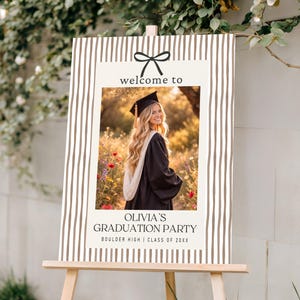 Pode incluir: Uma placa de festa de formatura em um cavalete de madeira. A placa tem uma foto de uma graduada em traje e chap&eacute;u, com o texto "welcome to" e "Olivia's Graduation Party". O fundo tem um padr&atilde;o listrado.