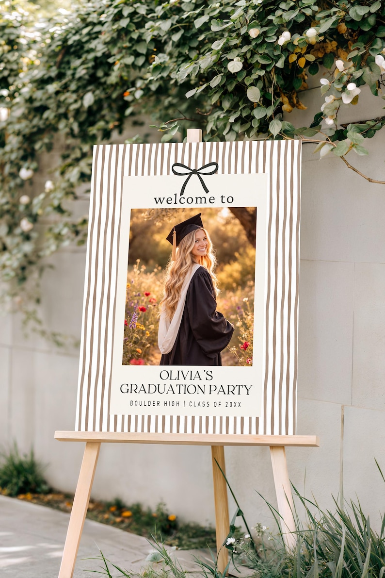 Pode incluir: Uma placa de festa de formatura em um cavalete de madeira. A placa tem uma foto de uma graduada em traje e chap&eacute;u, com o texto "welcome to" e "Olivia's Graduation Party". O fundo tem um padr&atilde;o listrado.