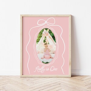 May include: A framed pink print with a white bow design at the top and a white oval outline. Inside the oval is a photo of a baby with a cake. The text "Holly is One" is written in white script at the bottom.