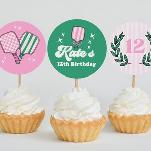 May include: Three cupcakes with white icing, each topped with a decorative circle. One pink circle features pickleball paddles, another green circle says "Kate's 12th Birthday", and the last has a pink "12" with a green wreath.