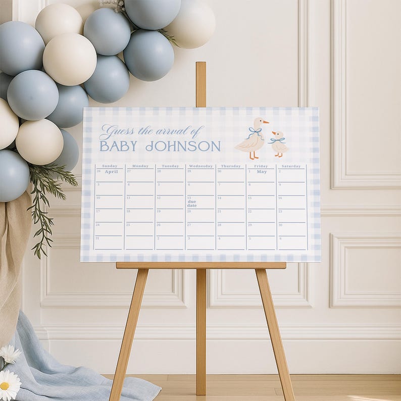 May include: A baby shower calendar with a blue gingham border and the text "Guess the arrival of BABY JOHNSON." The calendar features a duck illustration and a space to mark the due date. It is displayed on a wooden easel, with blue and white balloons in the background.