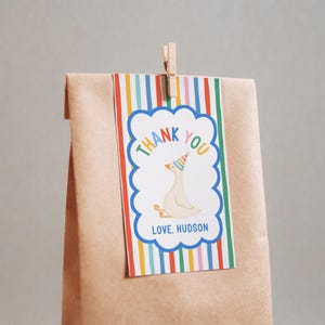 May include: A brown paper gift bag with a colorful striped tag. The tag features a cartoon goose wearing a party hat, the words "THANK YOU" in rainbow colors, and the name "HUDSON". A wooden clothespin holds the tag.