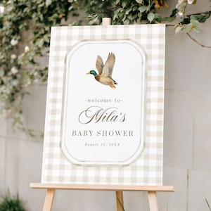 May include: A baby shower sign on a wooden easel. The sign has a light beige and white gingham pattern background. A duck illustration is in the center, above the text "Nila's BABY SHOWER August 15, 20XX".
