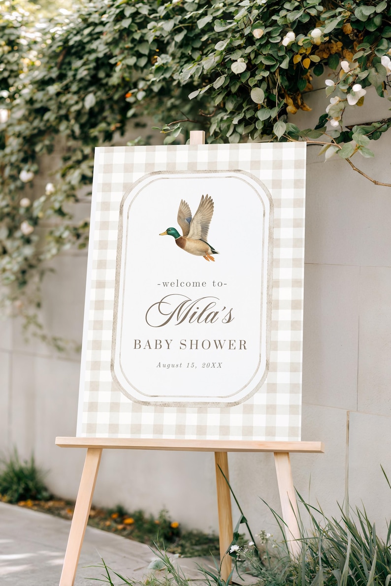 May include: A baby shower sign on a wooden easel. The sign has a light beige and white gingham pattern background. A duck illustration is in the center, above the text "Nila's BABY SHOWER August 15, 20XX".