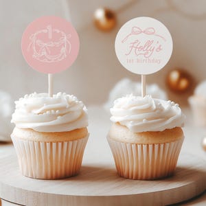 May include: Two cupcakes with white frosting and decorative toppers. One topper is pink with a cake illustration and the number 1. The other topper is white with the text "Holly's 1st Birthday" and a bow illustration. The cupcakes sit on a wooden board.