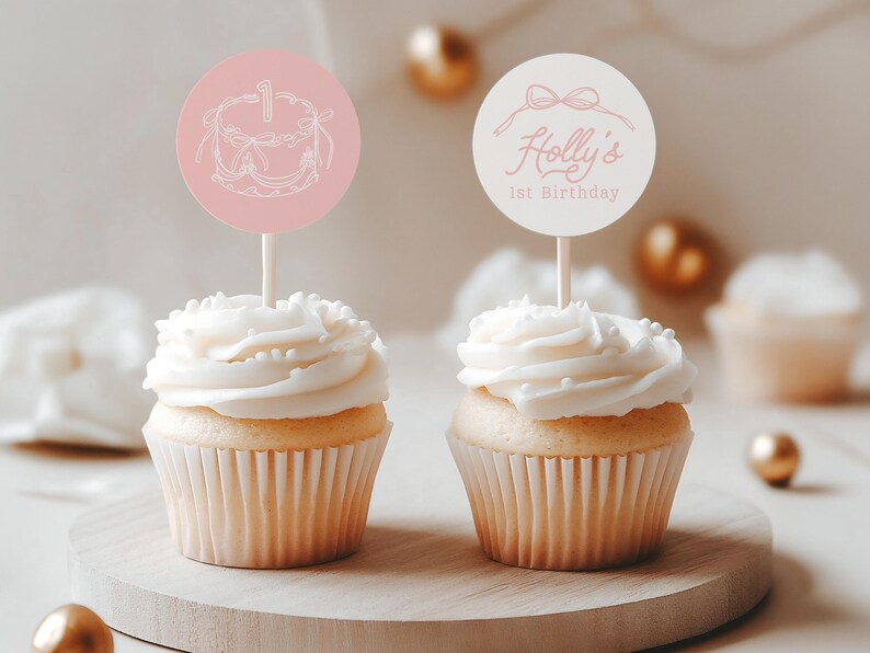 May include: Two cupcakes with white frosting and decorative toppers. One topper is pink with a cake illustration and the number 1. The other topper is white with the text "Holly's 1st Birthday" and a bow illustration. The cupcakes sit on a wooden board.