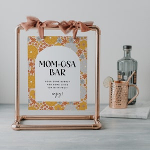 May include: A copper pipe stand with a sign that says "MOM-OSA BAR" with a floral pattern. The sign instructs to pour some bubbly, add some juice, and top with fruit. The sign also says "enjoy!"
