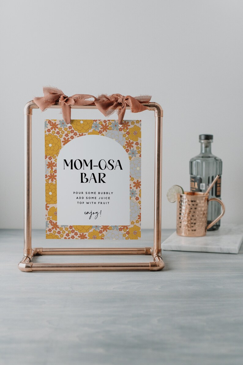 May include: A copper pipe stand with a sign that says "MOM-OSA BAR" with a floral pattern. The sign instructs to pour some bubbly, add some juice, and top with fruit. The sign also says "enjoy!"