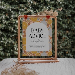 May include: A copper pipe stand with a sign that says "Baby Advice and Predictions". The sign has a floral pattern with yellow, orange, blue, and white flowers. The sign is on a white surface.