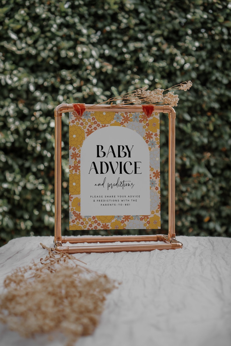 May include: A copper pipe stand with a sign that says "Baby Advice and Predictions". The sign has a floral pattern with yellow, orange, blue, and white flowers. The sign is on a white surface.