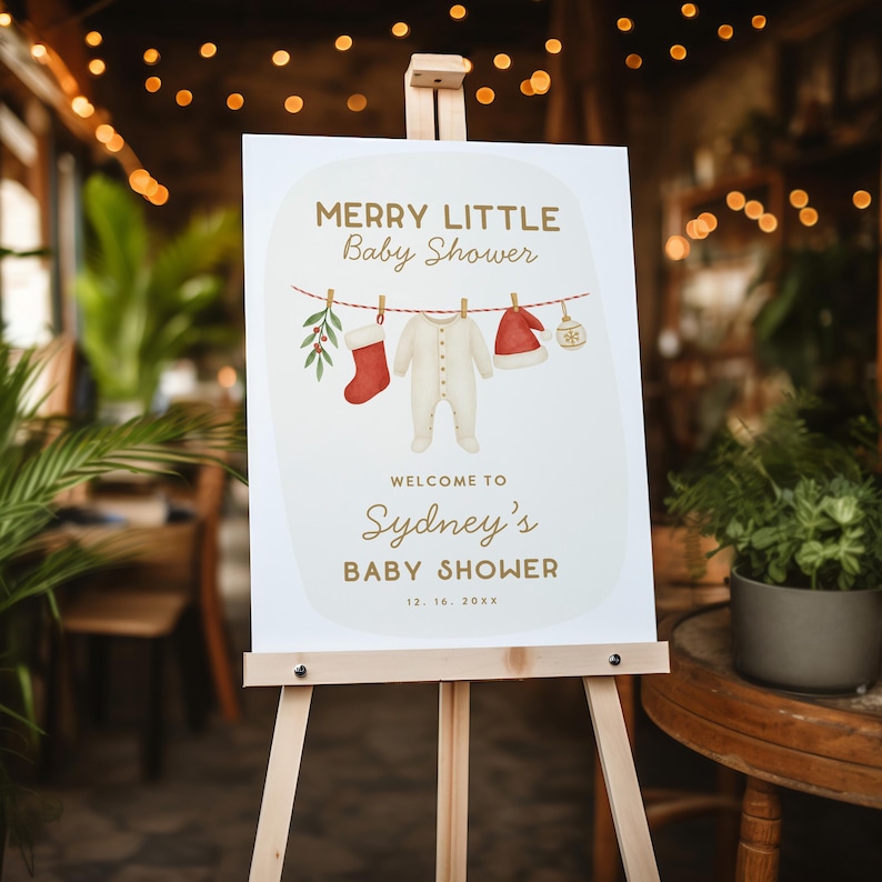 May include: A baby shower sign on a wooden easel. The sign reads "Merry Little Baby Shower" and "Welcome to Sydney's Baby Shower." The sign features illustrations of baby clothes and Christmas decorations. The date is 12.16.20XX.
