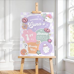 May include: A decorative sign on a wooden easel. The sign features a pastel color scheme with the words "Welcome to Luna's 1st Birthday Bash" and other text. The design includes smiley faces, ghosts, flowers, and stars.