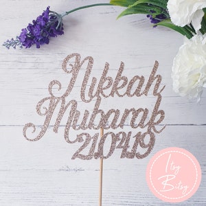 Può includere: Un topper per torta in glitter oro rosa con le parole "Nikkah Mubarak 2023" in un font corsivo. Il topper è su un bastoncino di legno ed è perfetto per una torta nuziale.