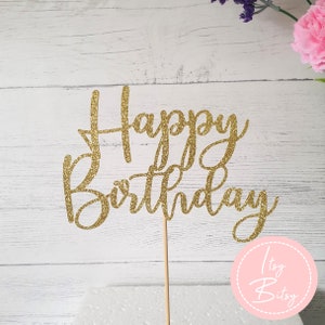 Puede incluir: Topper de pastel de purpurina dorada con las palabras "Happy Birthday" en una fuente cursiva. El topper está en un palo de madera y está sentado sobre una superficie blanca.