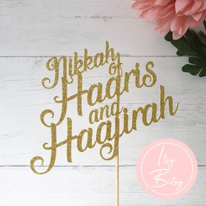 Può includere: Topper per torta con glitter dorati con le parole "Nikkah of Hagris and Hajirah" in un carattere corsivo. Il topper è attaccato a un bastoncino di legno. Un fiore rosa e un cerchio rosa con il testo "Itsy Bitsy" sono sullo sfondo.