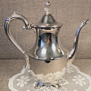 May include: A silver-plated coffee pot with a curved handle and spout. The pot has a decorative lid with a finial and sits on three ornate feet. The pot is highly reflective, showing the surrounding environment. The pot is placed on a white lace doily.