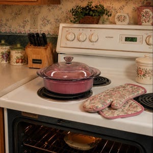 May include: A round, purple glass casserole dish with a matching lid, featuring vertical ribbed sides. The dish sits on a white electric stove burner next to a floral-patterned oven mitt. The stove has a digital clock displaying 12:18.