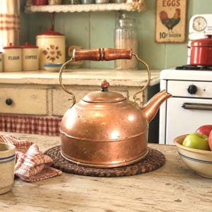 May include: A polished copper tea kettle with a rounded body, a curved spout, and a dark brown wooden handle. The kettle features a matching copper lid with a small knob and is positioned on a round, dark-toned woven mat on a wooden surface.