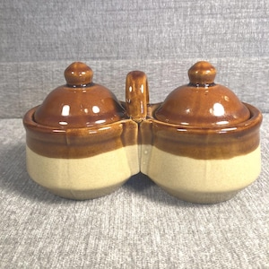 May include: A two-section ceramic condiment server with a handle. The server has a light beige base and is topped with a brown glaze. Each section has a matching lid with a small knob. The handle is also glazed brown.