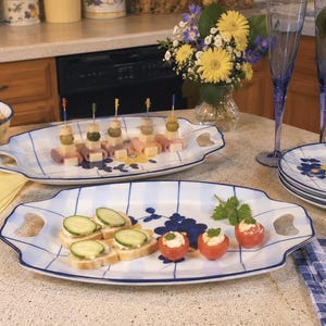 May include: Two white ceramic serving platters with blue grid patterns and yellow and blue floral accents. The platters feature integrated handles on each side. One platter holds appetizers on toothpicks, while the other holds cucumber slices on bread and stuffed tomatoes.