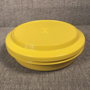 May include: A bright yellow, round food storage container with a matching lid. The container has a slightly flared base and a flat, circular lid with a small, embossed logo in the center. The container is made of a smooth, durable plastic.