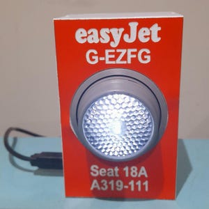 May include: A red box featuring the easyJet logo and the text "G-EZFG". A circular, silver light is in the centre, shining a bright white light. The text "Seat 18A A319-111" is at the bottom.